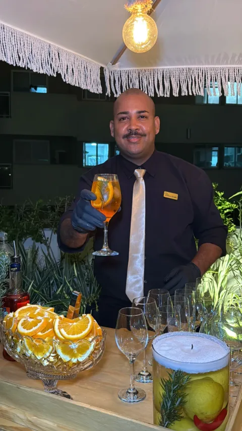 Bartender apresentando taça de Aperol Spritz em bar com frutas cítricas à noite